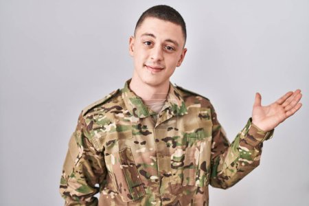 Téléchargez les photos : Jeune homme portant uniforme de camouflage de l'armée souriant gai présentation et pointant avec la paume de la main en regardant la caméra. - en image libre de droit