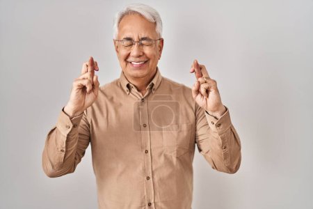 Photo for Hispanic senior man wearing glasses gesturing finger crossed smiling with hope and eyes closed. luck and superstitious concept. - Royalty Free Image