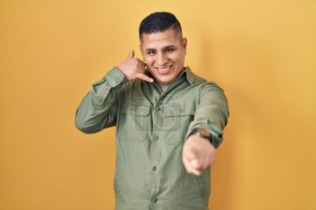 Téléchargez les photos : Jeune homme hispanique debout sur fond jaune souriant faire parler au téléphone geste et vous pointant du doigt. Appelle-moi. - en image libre de droit
