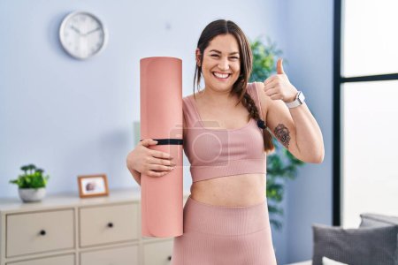 Téléchargez les photos : Jeune femme brune tenant tapis de yoga souriant heureux et positif, pouce levé faire excellent et signe d'approbation - en image libre de droit