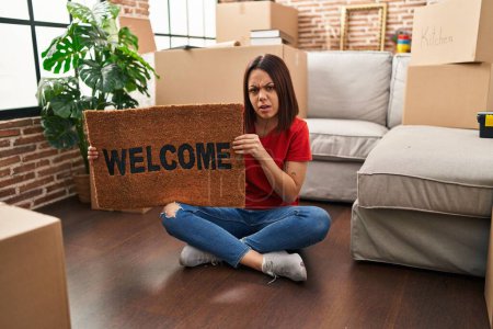 Photo for Young hispanic woman holding welcome doormat at new home clueless and confused expression. doubt concept. - Royalty Free Image