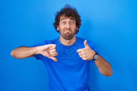 Photo for Hispanic young man standing over blue background doing thumbs up and down, disagreement and agreement expression. crazy conflict - Royalty Free Image