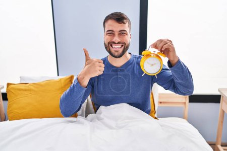 Foto de Hombre hispano guapo en la cama sosteniendo el despertador sonriendo feliz y positivo, pulgar hacia arriba haciendo excelente y señal de aprobación - Imagen libre de derechos