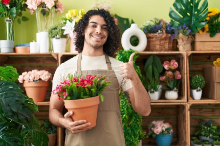 Téléchargez les photos : Homme hispanique avec les cheveux bouclés travaillant à la boutique de fleuriste tenant usine souriant heureux et positif, pouce levé faire excellent et signe d'approbation - en image libre de droit