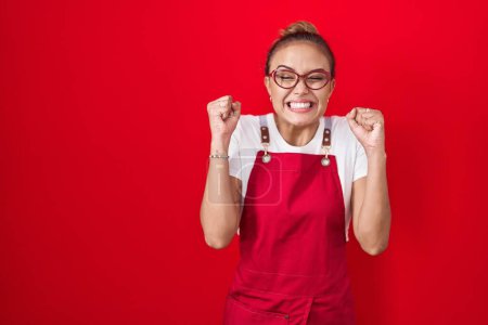 Téléchargez les photos : Jeune femme hispanique portant tablier serveuse sur fond rouge excité pour le succès avec les bras levés et les yeux fermés célébrant la victoire en souriant. concept gagnant. - en image libre de droit