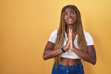 Téléchargez les photos : Femme afro-américaine avec les cheveux tressés debout sur fond jaune mendier et prier avec les mains ainsi que l'expression de l'espoir sur le visage très émotionnel et inquiet. mendier. - en image libre de droit
