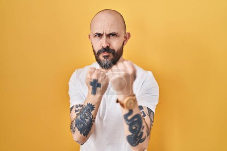 Foto de Joven hombre hispano con tatuajes de pie sobre fondo amarillo listo para luchar con el gesto de defensa del puño, cara enojada y molesta, miedo al problema - Imagen libre de derechos