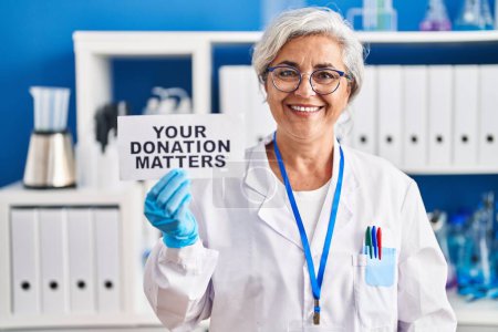 Foto de Mujer de mediana edad con el pelo gris trabajando en el laboratorio científico sosteniendo su pancarta de donaciones importa luciendo positiva y feliz de pie y sonriendo con una sonrisa confiada mostrando los dientes - Imagen libre de derechos