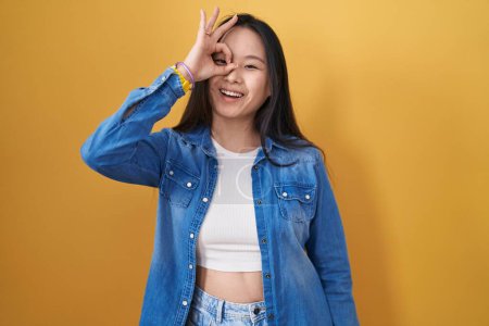 Foto de Joven asiático mujer de pie sobre amarillo fondo haciendo ok gesto con la mano sonriendo, ojo mirando a través de los dedos con feliz cara. - Imagen libre de derechos