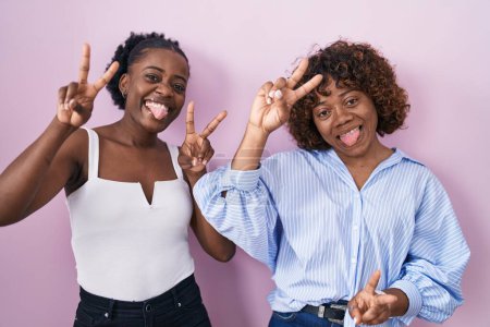 Téléchargez les photos : Deux femmes africaines debout sur fond rose souriant avec la langue montrant les doigts des deux mains faisant signe de victoire. numéro deux. - en image libre de droit