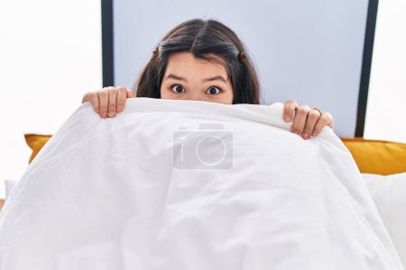 Téléchargez les photos : Jeune femme couvrant la bouche avec drap de lit assis sur le lit dans la chambre - en image libre de droit