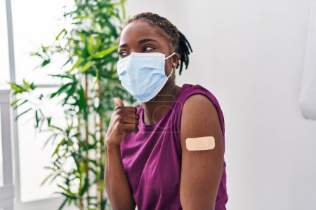 Photo for Beautiful black woman getting vaccine showing arm with band aid pointing thumb up to the side smiling happy with open mouth - Royalty Free Image