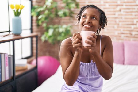 Foto de Mujer afroamericana de mediana edad bebiendo taza de café sentado en la cama en el dormitorio - Imagen libre de derechos