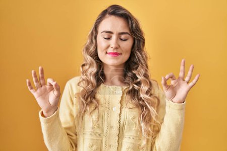 Téléchargez les photos : Jeune femme caucasienne debout sur fond jaune se détendre et sourire les yeux fermés en faisant geste de méditation avec les doigts. concept de yoga. - en image libre de droit