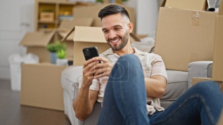 Photo for Young hispanic man using smartphone sitting on floor at new home - Royalty Free Image