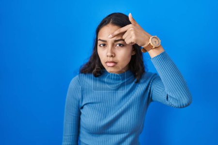 Téléchargez les photos : Jeune femme brésilienne debout sur fond bleu isolé pointant malheureux à boutons sur le front, infection laide de la tête noire. acné et problème de peau - en image libre de droit