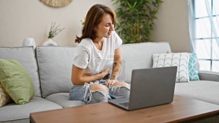 Téléchargez les photos : Jeune femme utilisant un ordinateur portable assis sur le canapé souffrant de maux d'estomac à la maison - en image libre de droit