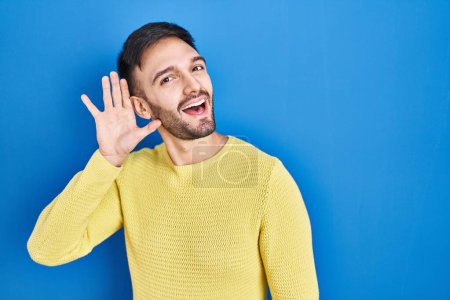 Téléchargez les photos : Homme hispanique debout sur fond bleu souriant avec la main sur l'oreille à l'écoute d'une audience à la rumeur ou des commérages. concept de surdité. - en image libre de droit