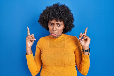Photo for Black woman with curly hair standing over blue background pointing up looking sad and upset, indicating direction with fingers, unhappy and depressed. - Royalty Free Image