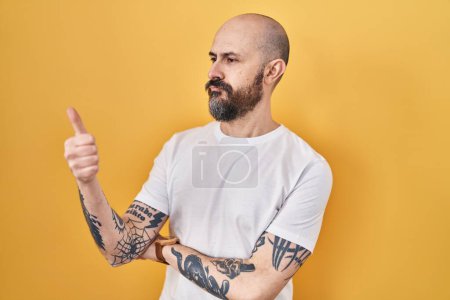 Foto de Joven hombre hispano con tatuajes de pie sobre fondo amarillo mirando orgulloso, sonriendo haciendo un gesto hacia un lado - Imagen libre de derechos