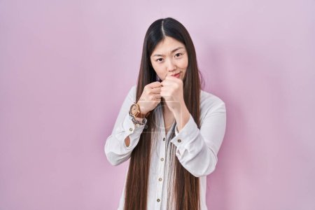 Téléchargez les photos : Jeune femme chinoise debout sur fond rose prêt à se battre avec geste de défense poing, visage en colère et bouleversé, peur du problème - en image libre de droit