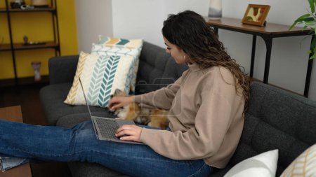 Photo for Young hispanic woman with dog using laptop sitting on sofa at home - Royalty Free Image
