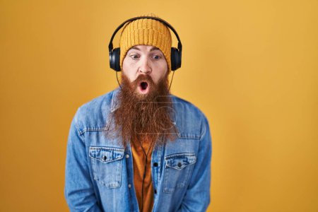 Téléchargez les photos : Homme caucasien à longue barbe écoutant de la musique à l'aide d'écouteurs dans le visage de choc, l'air sceptique et sarcastique, surpris avec la bouche ouverte - en image libre de droit