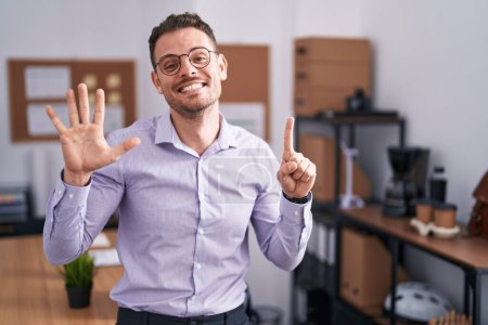 Foto de Joven hombre hispano en la oficina mostrando y señalando con los dedos número seis mientras sonríe confiado y feliz. - Imagen libre de derechos