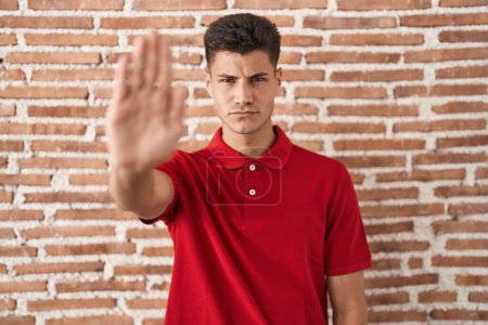 Téléchargez les photos : Jeune homme hispanique debout au-dessus du mur de briques faire arrêter chanter avec la paume de la main. expression d'avertissement avec geste négatif et sérieux sur le visage. - en image libre de droit