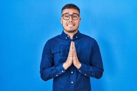 Téléchargez les photos : Jeune homme hispanique portant des lunettes sur fond bleu mendier et prier avec les mains ainsi que l'expression de l'espoir sur le visage très émotionnel et inquiet. mendier. - en image libre de droit