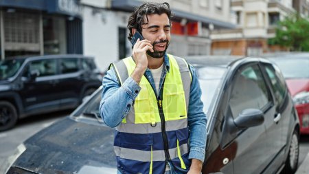 Téléchargez les photos : Jeune homme hispanique debout en voiture portant un gilet réfléchissant parlant sur smartphone dans la rue - en image libre de droit