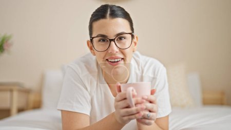 Téléchargez les photos : Jeune belle femme hispanique boire une tasse de café couché sur le lit dans la chambre - en image libre de droit