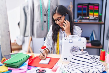 Photo for Young beautiful hispanic woman tailor talking on smartphone writing on notebook at clothing factory - Royalty Free Image