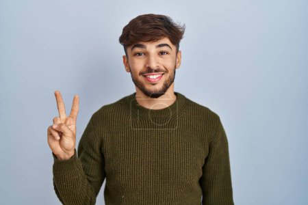 Téléchargez les photos : Homme arabe avec barbe debout sur fond bleu montrant et pointant vers le haut avec les doigts numéro deux tout en souriant confiant et heureux. - en image libre de droit