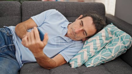 Téléchargez les photos : Homme d'âge moyen utilisant un smartphone couché sur le canapé à la maison - en image libre de droit