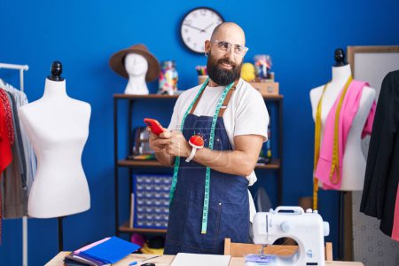 Téléchargez les photos : Jeune homme chauve tailleur souriant confiant en utilisant smartphone à l'usine de vêtements - en image libre de droit