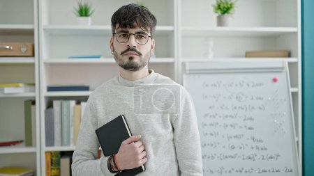 Photo for Young hispanic man teacher standing serious holding book at university classroom - Royalty Free Image