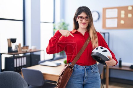 Photo for Young brunette woman working at the office holding bike helmet pointing finger to one self smiling happy and proud - Royalty Free Image