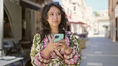 Téléchargez les photos : Jeune belle femme hispanique utilisant un smartphone avec une expression sérieuse sur la terrasse du café - en image libre de droit