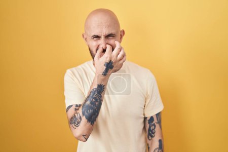 Téléchargez les photos : Homme hispanique avec des tatouages debout sur fond jaune sentant quelque chose de puant et dégoûtant, odeur intolérable, retenant souffle avec les doigts sur le nez. mauvaise odeur - en image libre de droit
