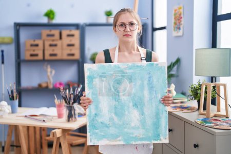Foto de Mujer caucásica joven sosteniendo lienzo en el estudio de arte escéptico y nervioso, frunciendo el ceño molesto debido a un problema. persona negativa. - Imagen libre de derechos