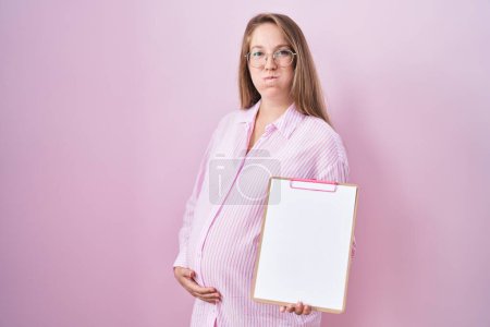 Foto de Mujer embarazada joven sosteniendo portapapeles soplando mejillas con cara divertida. boca inflada con aire, captación de aire. - Imagen libre de derechos