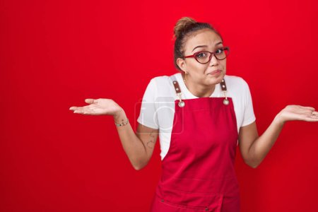 Photo for Young hispanic woman wearing waitress apron over red background clueless and confused expression with arms and hands raised. doubt concept. - Royalty Free Image