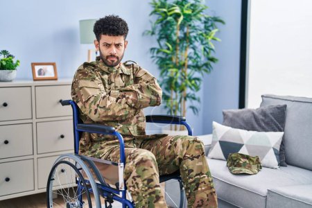 Photo for Arab man wearing camouflage army uniform sitting on wheelchair skeptic and nervous, disapproving expression on face with crossed arms. negative person. - Royalty Free Image
