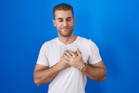 Photo for Young caucasian man standing over blue background smiling with hands on chest with closed eyes and grateful gesture on face. health concept. - Royalty Free Image