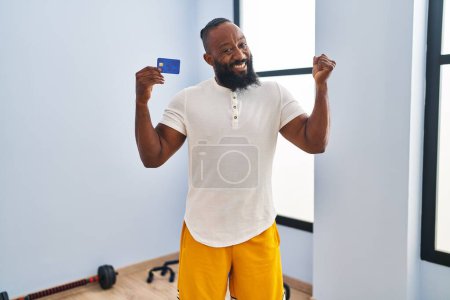 Foto de Hombre afroamericano usando ropa deportiva sosteniendo tarjeta de crédito gritando orgulloso, celebrando la victoria y el éxito muy emocionado con el brazo levantado - Imagen libre de derechos