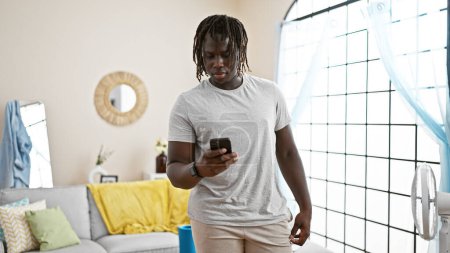 Foto de Hombre afroamericano de pie con cara seria usando smartphone en casa - Imagen libre de derechos