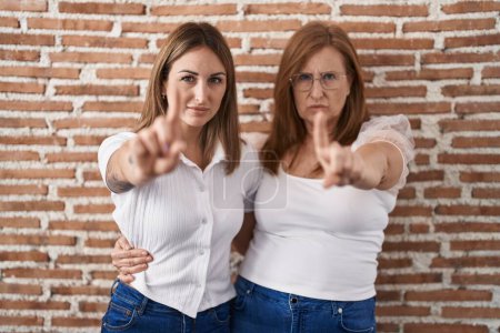 Téléchargez les photos : Mère et fille hispaniques portant un t-shirt blanc décontracté pointant le doigt vers le haut et l'expression en colère, ne montrant aucun geste - en image libre de droit