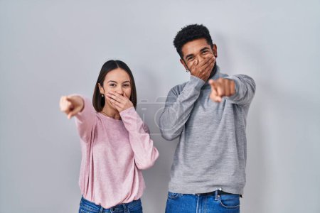 Foto de Joven pareja hispana de pie juntos riéndose de ti, señalando con el dedo a la cámara con la mano sobre la boca, expresión de vergüenza - Imagen libre de derechos