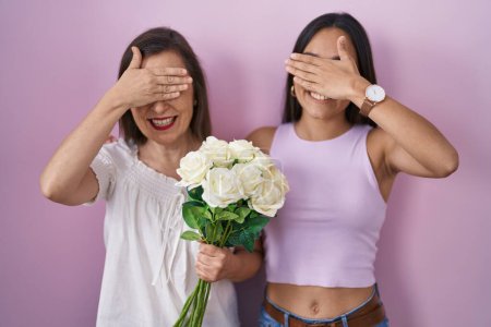 Téléchargez les photos : Mère et fille hispaniques tenant un bouquet de fleurs blanches souriant et riant avec la main sur le visage couvrant les yeux pour la surprise. concept aveugle. - en image libre de droit